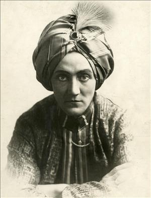 A seated portrait of a person wearing a turban on their head, dramatic eye makeup, and a loose tunic. They are staring intensely into the camera. 